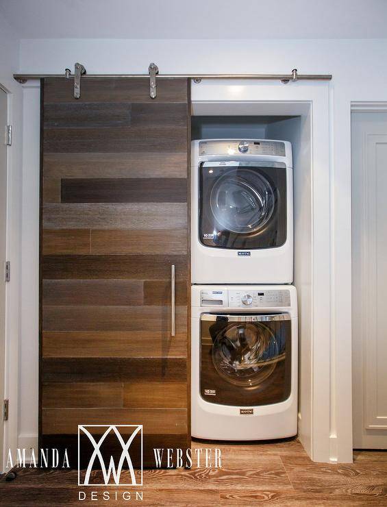 A stunning plank barn door on rails opens to reveal a small hall laundry room fitted with a stacked white front loading washer and dryer.