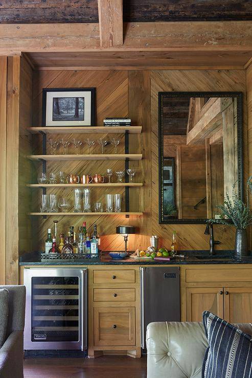 Country wet bar features iron and wood shelves mounted against a salvaged wood backsplash beside a black framed mirror. The mirror hangs above a sink with an oil rubbed bronze faucet kit fixed to a black marble countertop finishing salvaged wood cabinets. The cabinets are finished with oil rubbed bronze knobs and a stainless steel mini fridge and a glass front wine fridge.