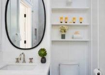 White stacked shelves over the toilet are featured in this charming toilet nook that boasts an oblong matte black mirror over a blue wooden single washstand with silver bamboo pulls.