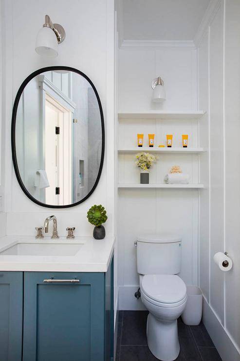 White stacked shelves over the toilet are featured in this charming toilet nook that boasts an oblong matte black mirror over a blue wooden single washstand with silver bamboo pulls.
