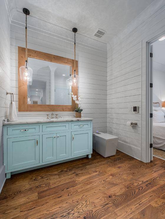 Rustic glass and rope lights over a turquoise blue bath vanity with a white marble top. White shiplap walls offer a cottage feel surrounding stained oak hardwood floors complimenting a wood framed vanity mirror.