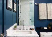 A two light bronze and glass sconce is mounted to a blue wall over a frameless vanity mirror hung above a small white single washstand. The washstand is accented with an oil rubbed bronze pull and a marble countertop holding a polished nickel gooseneck faucet in front of a marble backsplash.