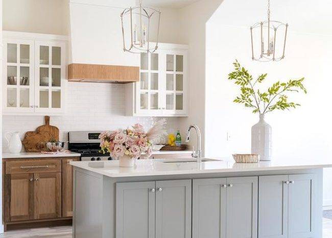 Light stained wood floors frame a gray kitchen island lit by two French nickel lanterns. Stained wood cabinets are topped with a white countertop fixed against glossy white staggred backsplash tiles beneath glass front cabinets flanking a white range hood with wood trim.