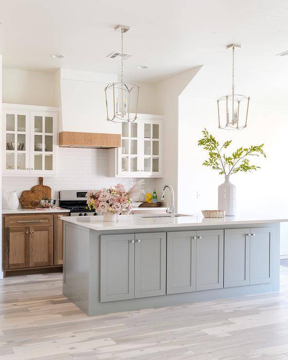 Light stained wood floors frame a gray kitchen island lit by two French nickel lanterns. Stained wood cabinets are topped with a white countertop fixed against glossy white staggred backsplash tiles beneath glass front cabinets flanking a white range hood with wood trim.