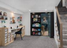 Basement craft room boasts black bookcases that swing open to reveal a movie room, and a black desk chair placed on gray carpeting a two-toned desk finished with white pull out bins lit by blue sconces. White floating shelves flank a pin board.
