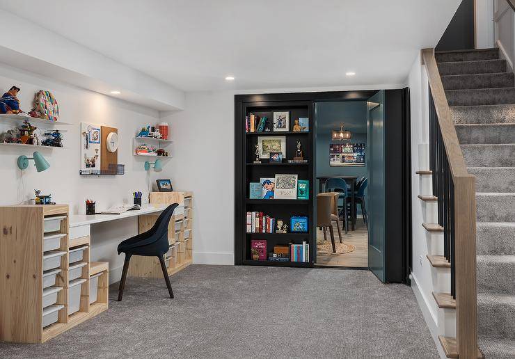 Basement craft room boasts black bookcases that swing open to reveal a movie room, and a black desk chair placed on gray carpeting a two-toned desk finished with white pull out bins lit by blue sconces. White floating shelves flank a pin board.