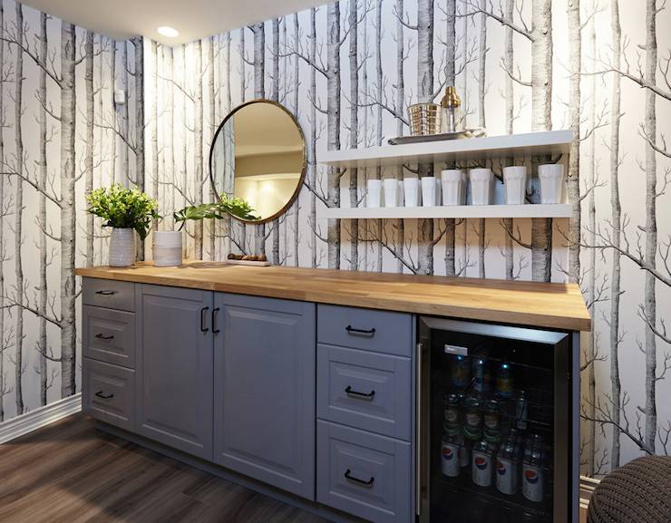 basement family room features bar fitted with gray cabinets and glass-front beverage fridge topped with butcher block under round mirror next to stacked floating shelves mounted on wall covered in Cole & Sons Woods Wallpaper.