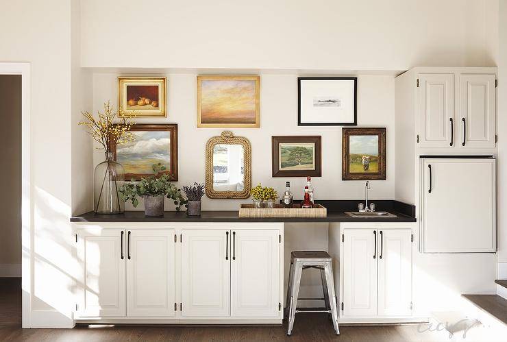 Basement wet bar features white raised panel cabinets topped with black countertops and a West Elm Raffia Tray filled with libations next to a small bar sink situated under an eclectic collection of art and a rope mirror beside an under cabinet fridge alongside a Tolix Stool.