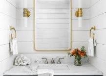 A shiny brass mirror on a shiplap wall hangs over a black shaker washstand adorned with brass drop pulls.