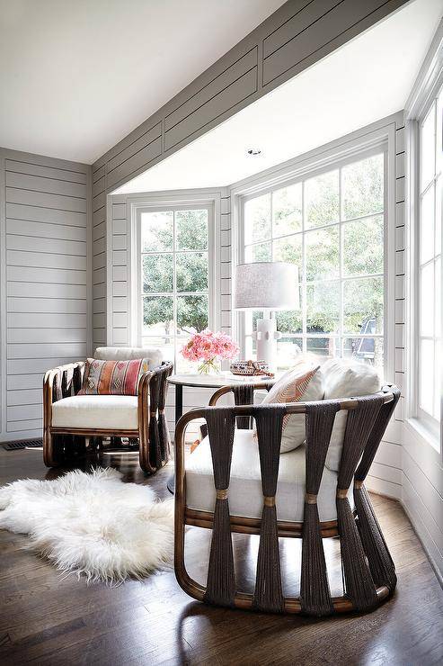 A white sheepksin rug sits in front of gray rope and wood lounge chairs placed on a stained wood floor in front of pay windows framed by gray shiplap.