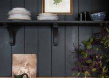 A black kitchen shelf is mounted to a black shiplap wall beneath a framed botanical art piece and over a wooden countertop.