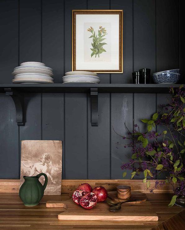 A black kitchen shelf is mounted to a black shiplap wall beneath a framed botanical art piece and over a wooden countertop.