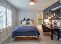 Boy's room boasts walls clad in white and blue zigzag wallpaper lined with a stained wood bed dressed in a green sheet set and a blue border duvet and shams situated across from a dark stained desk paired with matching colored chair situated under a collage of black and white football photos mural.