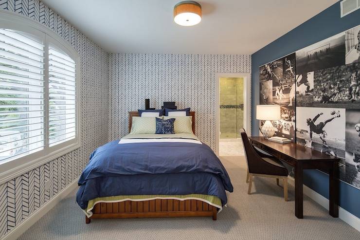 Boy's room boasts walls clad in white and blue zigzag wallpaper lined with a stained wood bed dressed in a green sheet set and a blue border duvet and shams situated across from a dark stained desk paired with matching colored chair situated under a collage of black and white football photos mural.