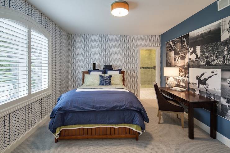 Boy's room boasts walls clad in white and blue zigzag wallpaper lined with a stained wood bed dressed in a green sheet set and a blue border duvet and shams situated across from a dark stained desk paired with matching colored chair situated under a collage wall of black and white football photos mural.