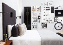 Chic black and white contemporary bedroom boasts a tall black headboard lit by black plug in sconces and accenting a bed dressed in white and gray bedding topped with gray and black pillows. A tall black armoire is located beside a gorgeous black and white gallery art wall.