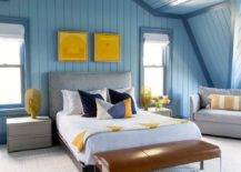 Boy's room features a gray velvet headboard with yellow and blue bedding on vertical blue shiplap trim, gray modern nightstands, a brown leather and lucite bench at the foot of the bed, a blue shiplap vaulted ceiling and a tan sofa.
