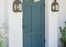 Blue wooden chevron gate door completed with oil rubbed bronze hardware illuminated by vintage sconces flanked by a white brick wall.