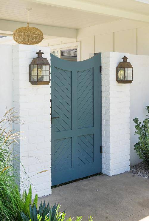 Blue wooden chevron gate door completed with oil rubbed bronze hardware illuminated by vintage sconces flanked by a white brick wall.