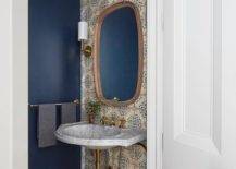 Small, luxurious powder room is styled with an oblong mirror hung from a wall clad in cream and blue mosaic wall tiles between brass sconces and over a French marble wall mount sink with an antique brass cross handle faucet.