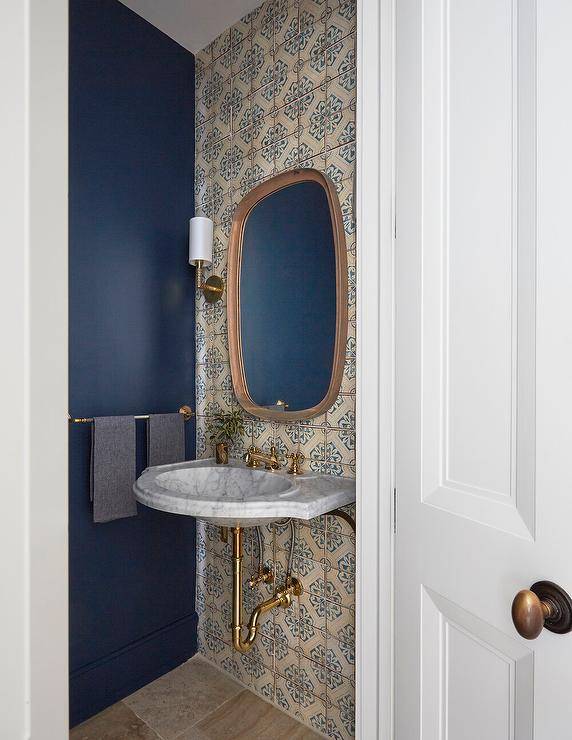 Small, luxurious powder room is styled with an oblong mirror hung from a wall clad in cream and blue mosaic wall tiles between brass sconces and over a French marble wall mount sink with an antique brass cross handle faucet.