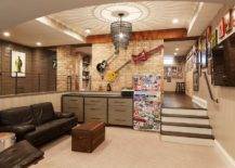 Well appointed sunken man cave features a black chain tiered chandelier illuminating a black leather sofa placed facing a trunk coffee table and a wall mount television mounted beneath framed art. This basement is also fitted with a sticker covered mini fridge placed beside a two toned black and gray dresser as decorative guitars are mounted above it on a brick wall.