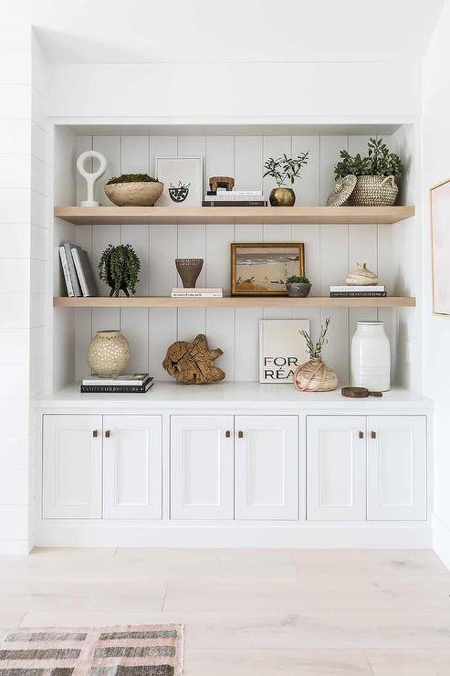 Gray wash floating shelves sit on white vertical shiplap trim over white built in cabinets accented with brass pulls in a bedroom.