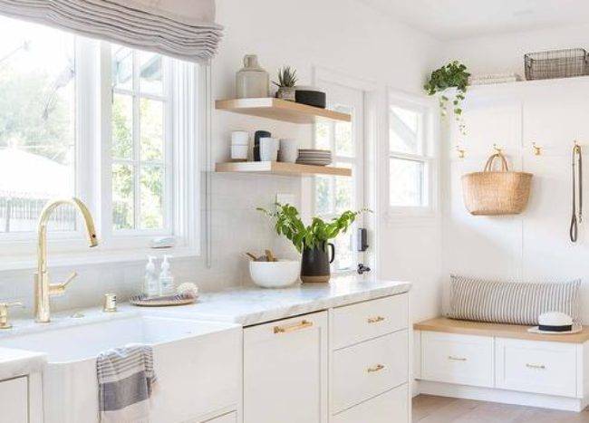 Off to the side of a lovely white kitchen, a wood top storage bench is accented with a gray striped lumbar pillow and placed against a wainscot backsplash and beneath brass hooks mounted under a white shelf.