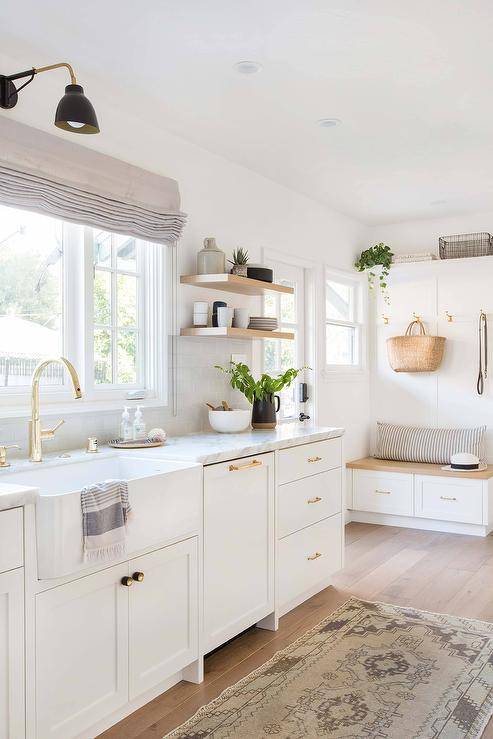 Off to the side of a lovely white kitchen, a wood top storage bench is accented with a gray striped lumbar pillow and placed against a wainscot backsplash and beneath brass hooks mounted under a white shelf.