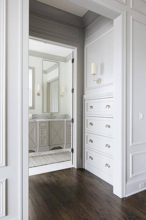 A Ziyi Sconce mounted to a gray wainscot wall illuminates light gray built-in hallway drawers accented with polished nickel cup pulls. The hallway leads to an en suite master bathroom.