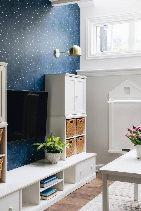 A blue and white basement playroom features dark blue stars wallpaper, illuminated by a brass sconce, along with a white coffee table atop a white and taupe rug. Underneath the window is a white wall displaying a memo board. The space is completed with a built-in white tv media cabinet, complete with a built in toy storage case and taupe woven bins, encasing an attached tv.