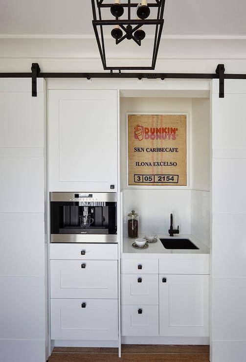 Cottage kitchen features a built-in coffee station hidden by sliding barn doors on rails. A vintage Dunkin donuts sign decorates the built-in finished with a sink, an oil rubbed bronze faucet, a wold built-in coffee machine, and white shaker cabinets. Oil rubbed bronze hardware stand out against the white finished cabinetry keeping a black and white theme consistent.