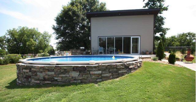 above ground pool with stone wall built into a hill with house in background on grass