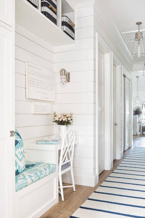 A blue stripe rug lines a cottage hallway boasting a white built-in desk matched with a white bamboo desk chair and positioned beside a white built-in bench with turquoise blue ikat cushions. The desk is fixed beneath stacked art mounted to a white tongue and groove wall beneath white overhead cubbies and lit by a cage sconce.