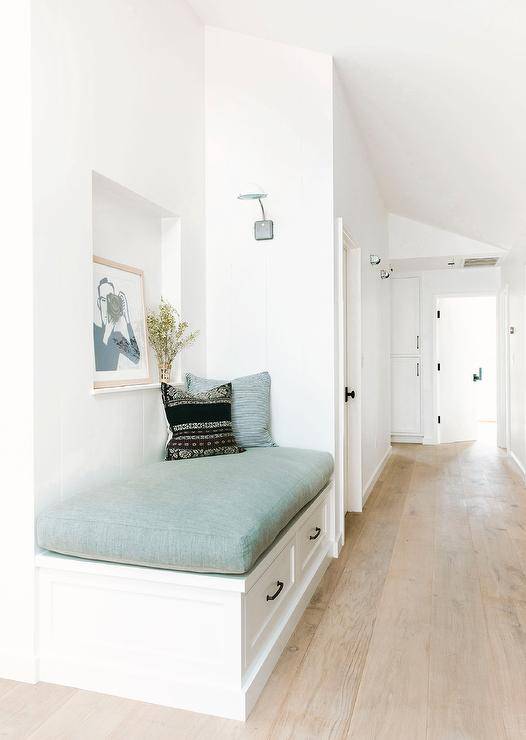 A white built-in foyer reading bench is fitted with drawers donning oil rubbed bronze pulls and topped with a blue seat cushion. The bench is positioned beneath a niche holding an art piece.