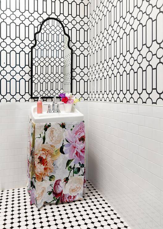 This chic white and black contemporary powder room is accented with a small floral skirted sink vanity finished with a polished nickel faucet kit and fixed under a black arch mirror. Walls are half clad in white and black trellis wallpaper and finished with white subway tiles complementing white and black vintage floor tiles.