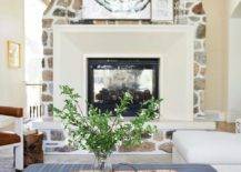 Living room features a limestone and stone double sided fireplace design and a woven tray on a blue striped ottoman used as a coffee table.