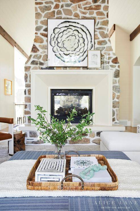 Living room features a limestone and stone double sided fireplace design and a woven tray on a blue striped ottoman used as a coffee table.