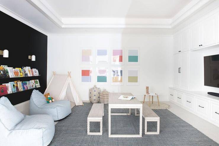 Amazing contemporary playroom is fitted with wall-to-wall black built-in bookcases fixed behind light gray modern bean bags placed on a black and white grid rug beside a teepee. Colorful abstract art hangs over Moroccan style toy baskets, while a desk is matched with benches and locatd under a tray ceiling.