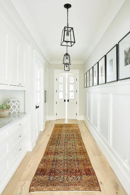 Seeded glass lanterns hang over a vintage runner placed on a blond wood floor leading to arched double doors contrasted with oil rubbed bronze knobs. A wall half fitted with a board and batten trim is lined with a black and white photo gallery hung facing white cabinets fixed over white drawers.