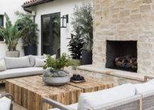 Patio covered by a white fabric pergola features wood and gray rope outdoor sofas with a reclaimed wood coffee table, a stone fireplace and hearth, and potted olive trees.