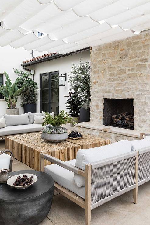 Patio covered by a white fabric pergola features wood and gray rope outdoor sofas with a reclaimed wood coffee table, a stone fireplace and hearth, and potted olive trees.