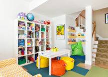 Basement playroom features an under the stairs reading nook lined with floating bookshelves and a neon green bean bag. Playroom in basement boasts an Ikea Expedit Shelving Unit filled with book as and toys alongside a white play table lined with yellow and orange poufs atop blue and green carpet tiles across from a white and yellow chevron sofa.