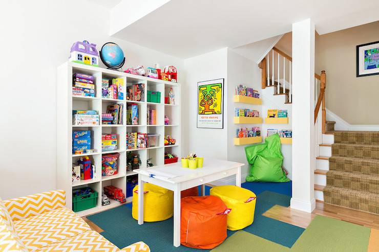 Basement playroom features an under the stairs reading nook lined with floating bookshelves and a neon green bean bag. Playroom in basement boasts an Ikea Expedit Shelving Unit filled with book as and toys alongside a white play table lined with yellow and orange poufs atop blue and green carpet tiles across from a white and yellow chevron sofa.