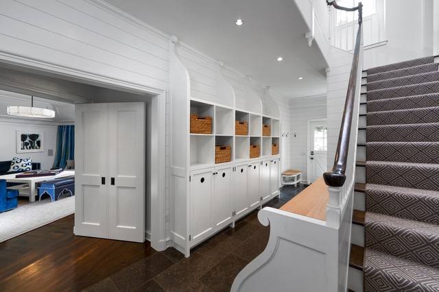 Fabulous entryway with white paneled walls and tiled floors bordering hardwood floors in the family room. The entryway features built-in mudroom storage with closet cabinets and open cubbies lined with woven storage bins. A Dutch door at the end of the hall leads to the garden. The stairway is finished with a gray and white Greek Key stair runner.