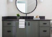 Oil rubbed bronze and white glass sconces flank a round black mirror hung over a gray industrial washstand adorned with oil rubbed bronze hardware and an oil rubbed bronze faucet mounted to a black quartz countertop. The washstand sits on small black hexagon floor tiles.
