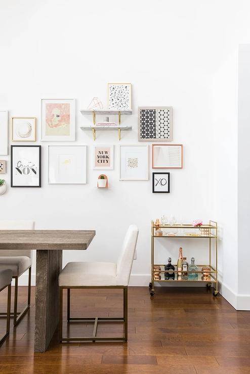 White and gold dining chairs surround a dark brown salvaged wood dining table placed in front of a gallery art wall located over a mirrored gold bar cart.