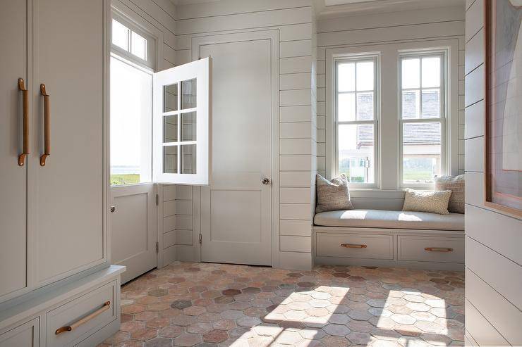A light gray Dutch door opens to a mudroom boasting terracotta hexagon floor tiles, light gray shiplap walls and a light gray built-in window seat accented with wood and leather pulls and a light gray cushion.