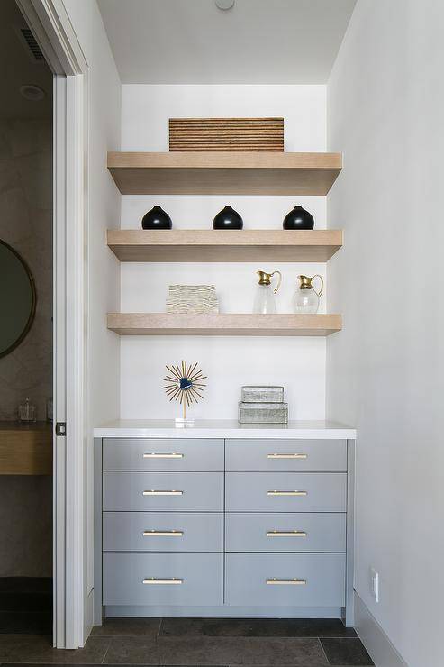 Bathroom hallway is fitted with gray cabinets adorning brass pulls and a white countertop fixed beneath three styled and stacked gray wash floating shelves.