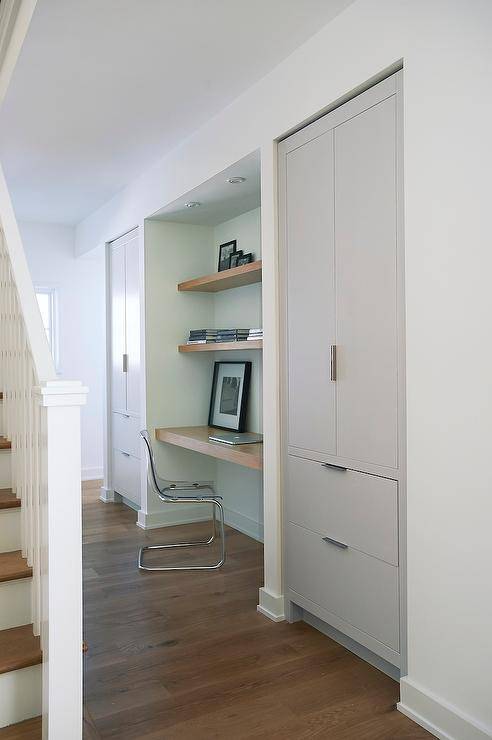 Foyer hall alcove is filled with a wood floating desk under stacked wood floating shelves paired with an Ikea Ghost Chair, Ikea Tobias Chair, flanked by alcoves filled with gray cabinets.